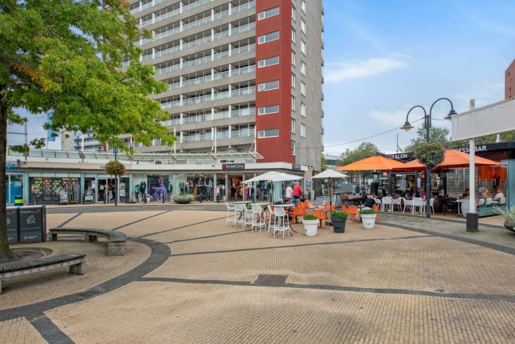 Terras met oranje parasols en winkels aan de Koningin Julianalaan in een stadsplein met hoogbouw.